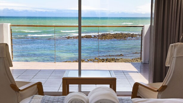 A hotel room with a bed, two chairs, a small table, and a view of the ocean through large windows. - Villa Kyma - Six Bedroom House, Sleeps 12 (Cape Town)