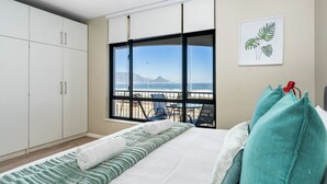 A bedroom with a bed, a window with a view of the beach, a balcony with chairs, and a white wardrobe.