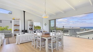 Un comedor moderno con una mesa de madera y sillas blancas, ventanas amplias que dan a una vista del mar, y una zona de estar acogedora con un sofá a cuadros.