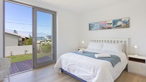 Un dormitorio con un amplio ventanal corredero que da a un balcón, una cama con ropa blanca, una mesita de noche con una lámpara y una obra de arte colgada en la pared.