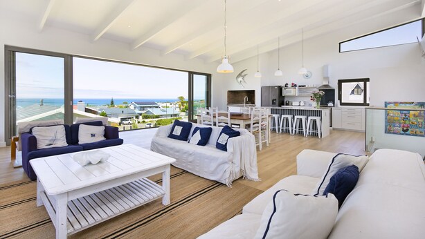 Amplia sala de estar con un ventanal que ofrece vistas al mar, una cocina con electrodomésticos modernos y un comedor con mobiliario blanco.