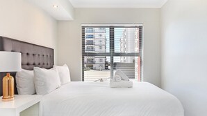 Ein Hotelzimmer mit einem großen Bett, einem Nachttisch mit Lampe und Blick auf die Stadt durch das Fenster.