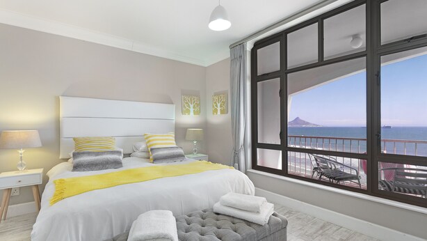 Ein Schlafzimmer mit großem Fenster, einem Bett mit weißer Bettwäsche, einer grau gepolsterten Bank und einem Nachttisch mit Lampe.