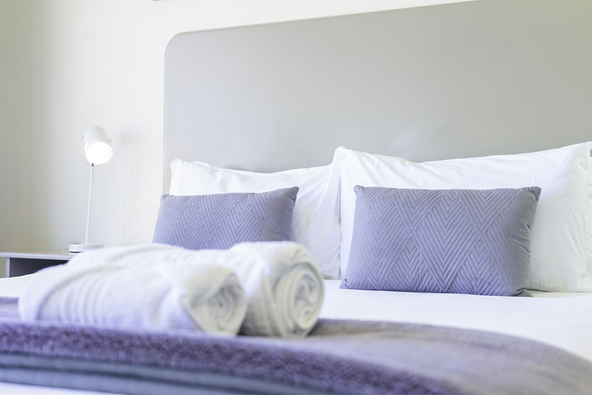 Una cama bien hecha con toallas blancas enrolladas, un cabecero blanco y dos almohadas con textura.