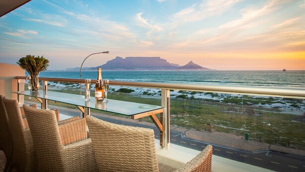 A balcony with wicker chairs and a glass table, overlooking a coastal view with a mountain in the distance. - Sea Spray B306 - Two Bedroom Apartment, Sleeps 4 (Cape Town)
