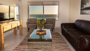 Ein Wohnzimmer mit großem Fenster, einem braunen Ledersofa, zwei Sesseln, einem Glastisch mit einer Vase und einer Holz-TV-Bank.