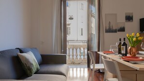 En moderne stue med en sofa, et spisebord med stole, udsigt til gaden fra en balkon, og en vase med blomster på bordet.