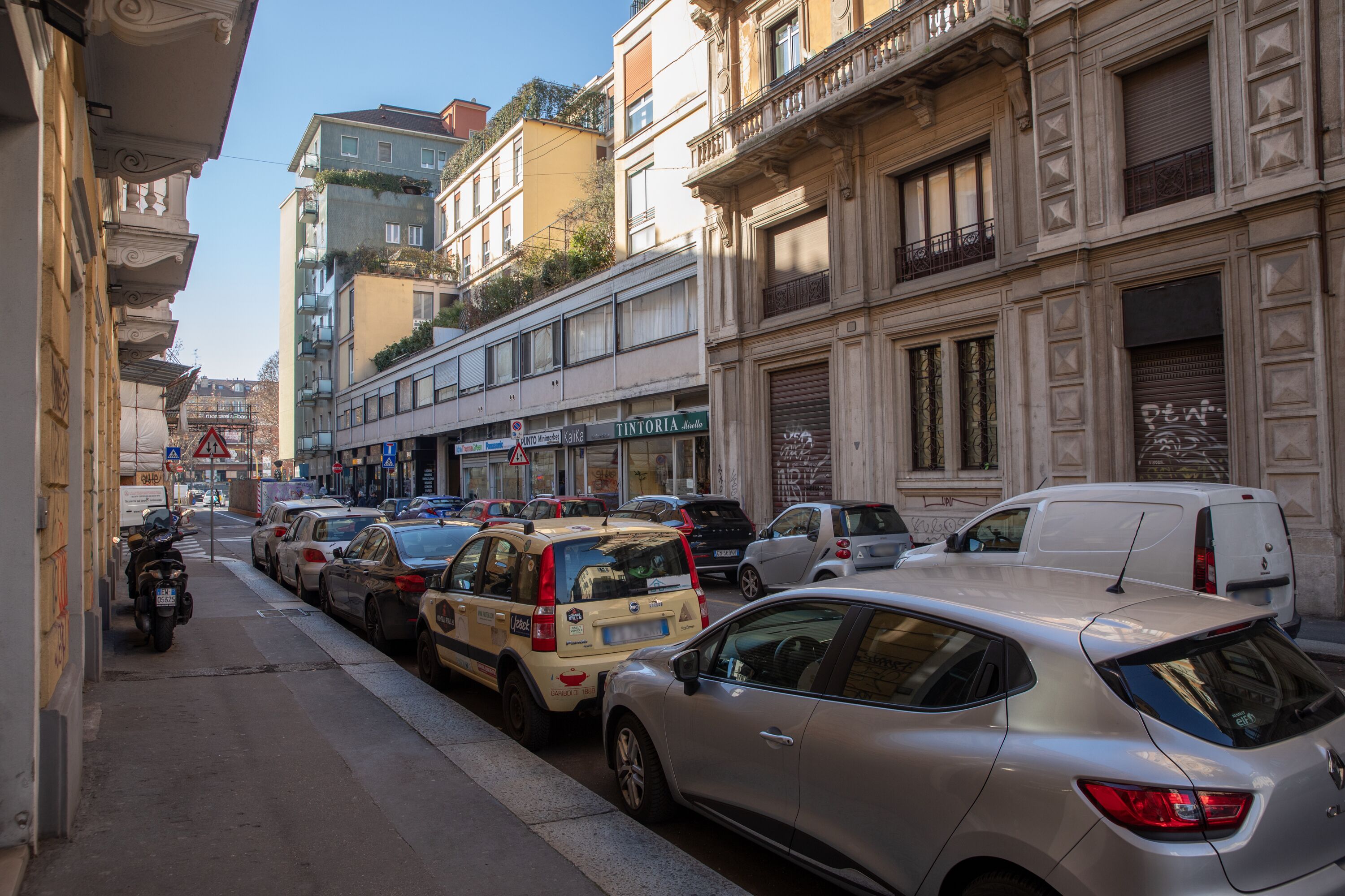 A narrow street lined with parked cars and buildings with balconies.