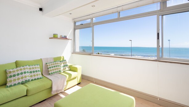 Een lichte kamer met een groene bank, uitzicht op zee en een raam met zonwering.