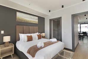 Quarto de hotel moderno com uma cama grande, cabeceira, mesa de cabeceira, abajur e um quadro na parede.