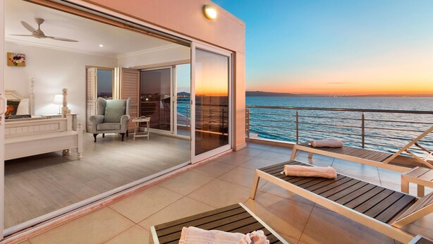 Un balcón con vistas al océano al atardecer, una zona de estar con una mesa y sillas, y una habitación con cama y sillón.