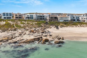 Eine Küstenlandschaft mit Sandstrand, felsiger Uferlinie und einer Reihe moderner Häuser mit einer Treppe, die zu einer höheren Lage führt.