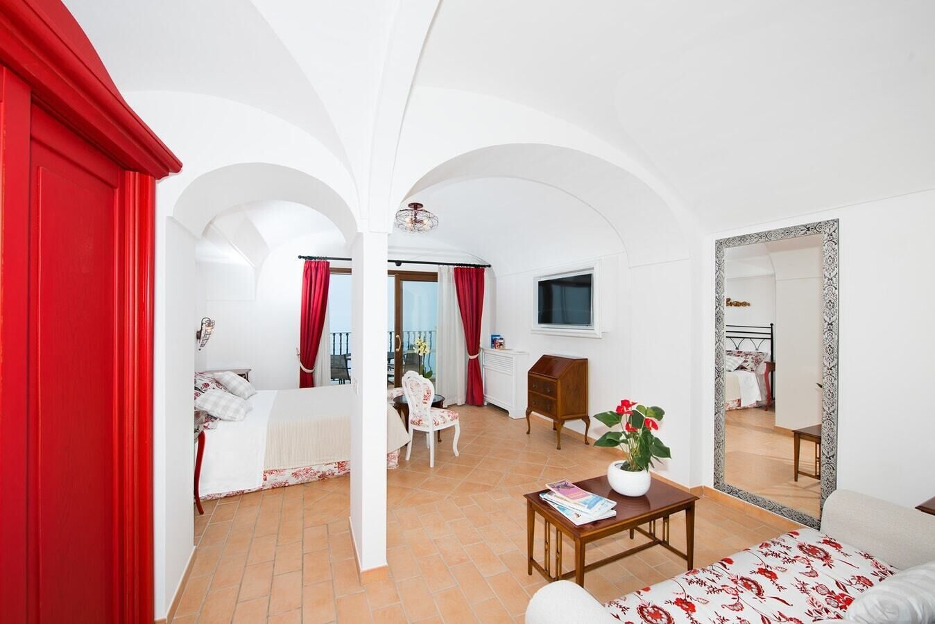 A hotel room with a bed, a small table with a plant, a TV, and a balcony with red curtains.