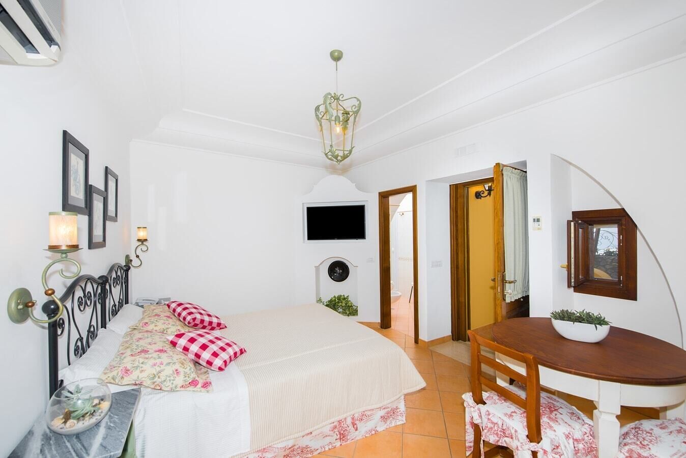 A bedroom with a bed, bedside table, lamp, wall-mounted TV, and a small table with a plant.