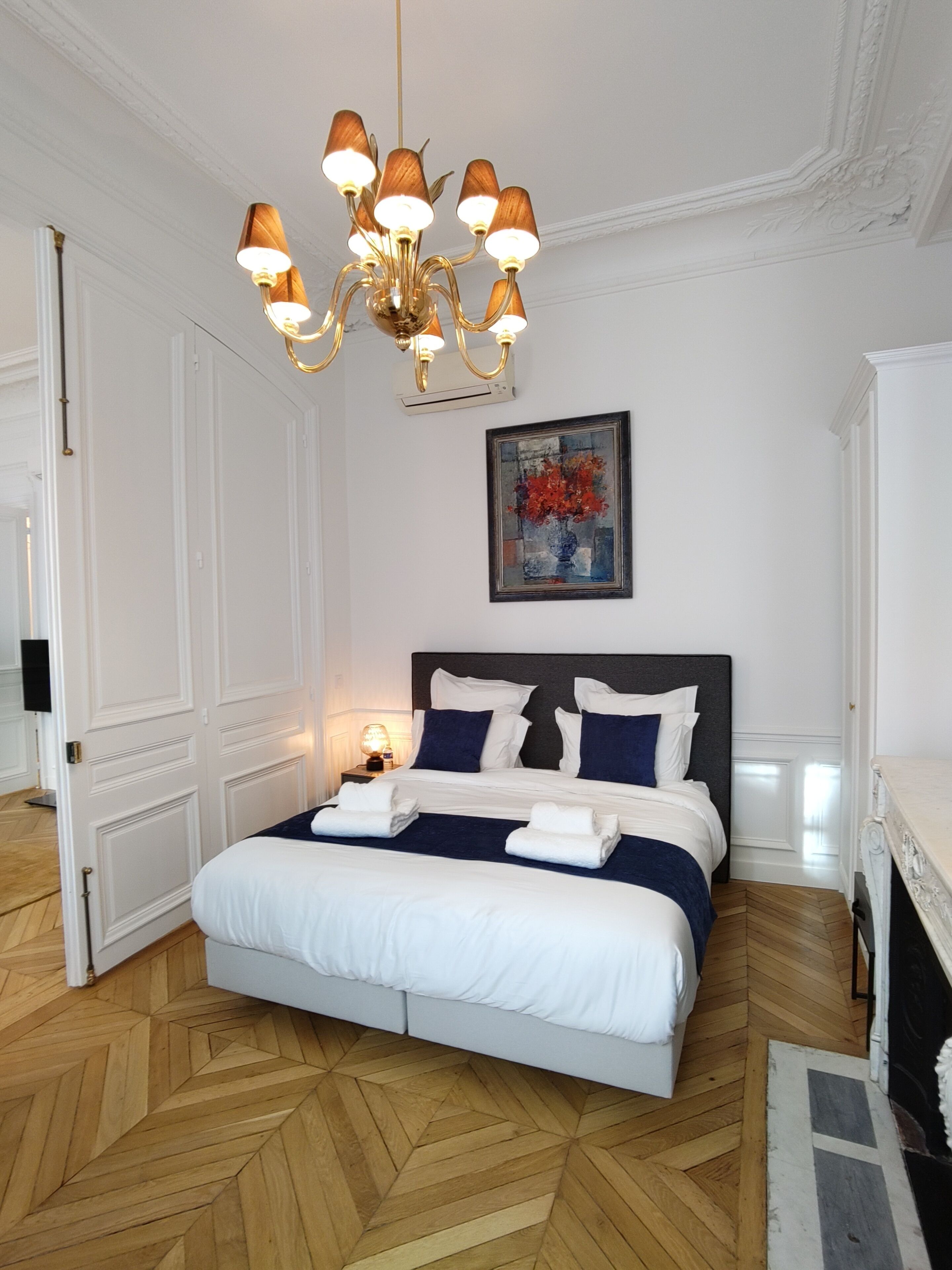 A hotel room with a large bed, a chandelier, a painting, and a fireplace.