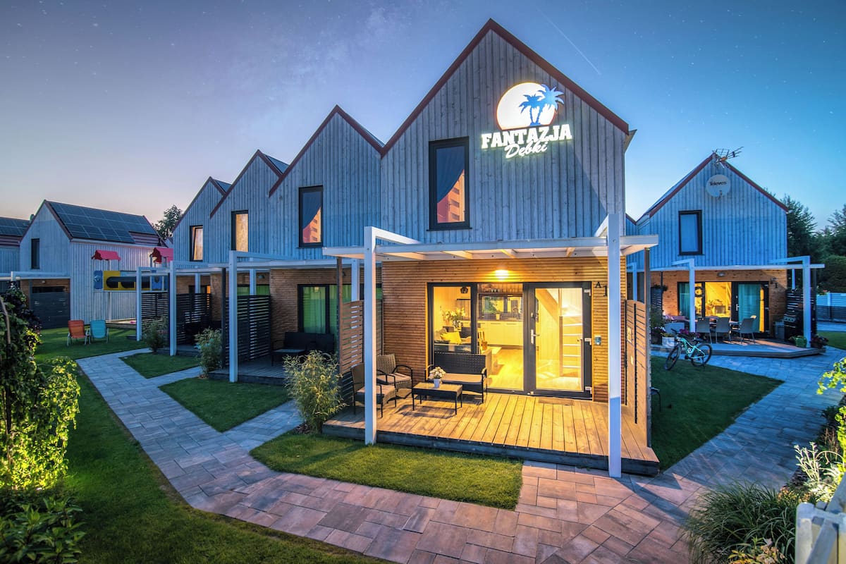 A modern building with a sign reading "Fantazija." It features a covered outdoor seating area with a table and chairs, and a well-maintained lawn.
