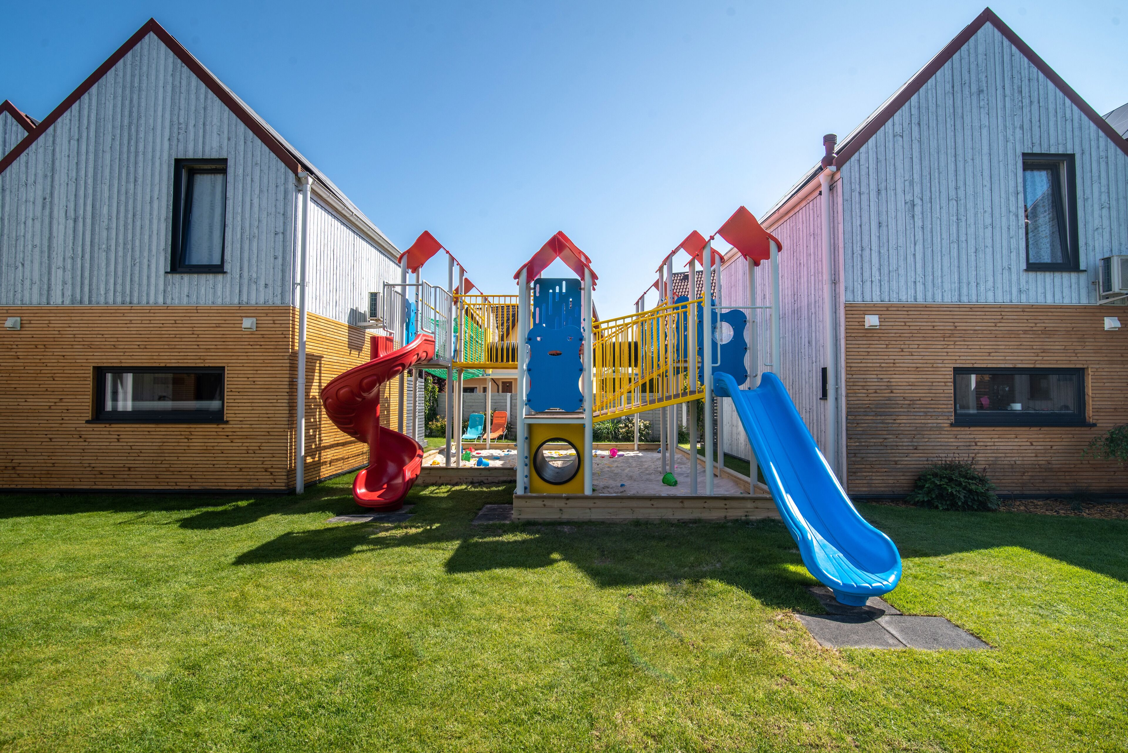 A playground with two slides and a climbing structure, set against a backdrop of wooden buildings.