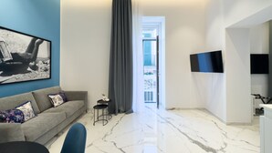 A modern living room with a grey sofa, a small black table, and a blue chair. There is a large window with a grey curtain, a flat-screen TV mounted on the wall, and a marble floor. - InSogno Suites (Naples)