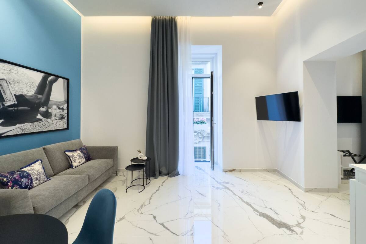 A modern living room with a grey sofa, a small black table, and a blue chair. There is a large window with a grey curtain, a flat-screen TV mounted on the wall, and a marble floor.