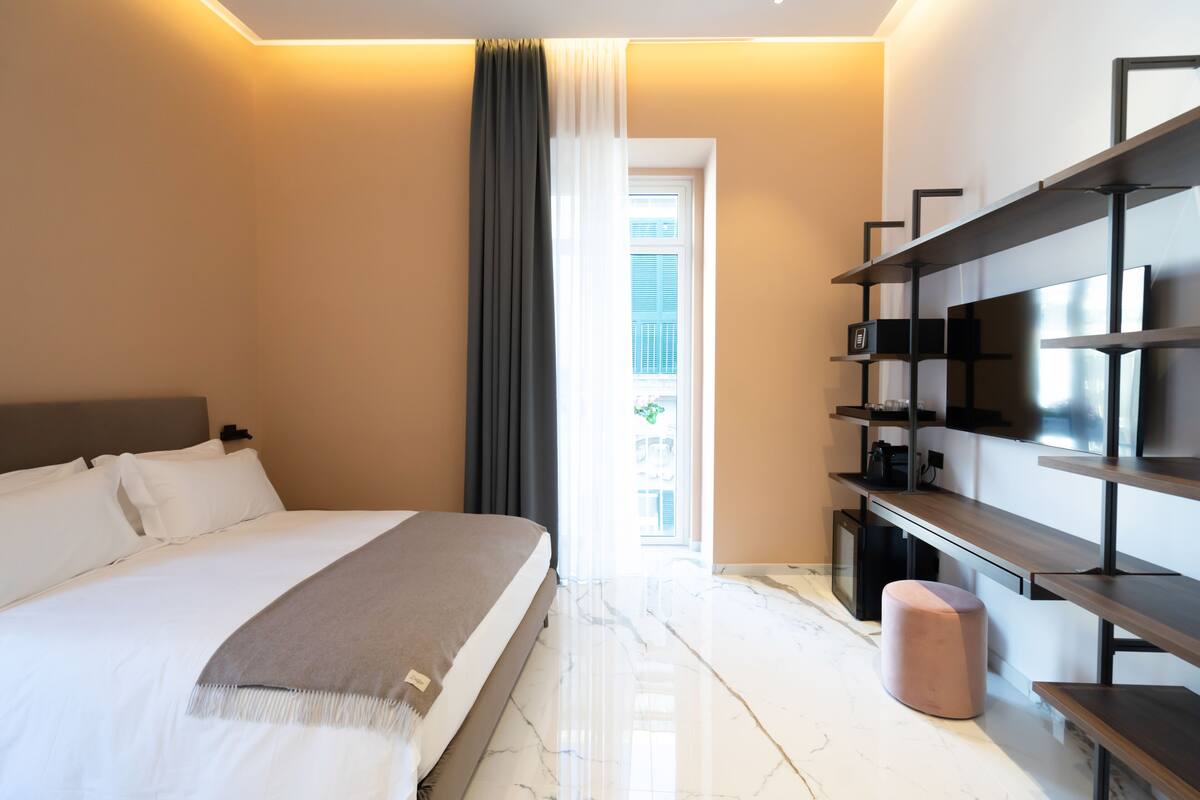 A modern hotel room with a large bed, a bedside table, a built-in shelf unit, and a view of the outside.