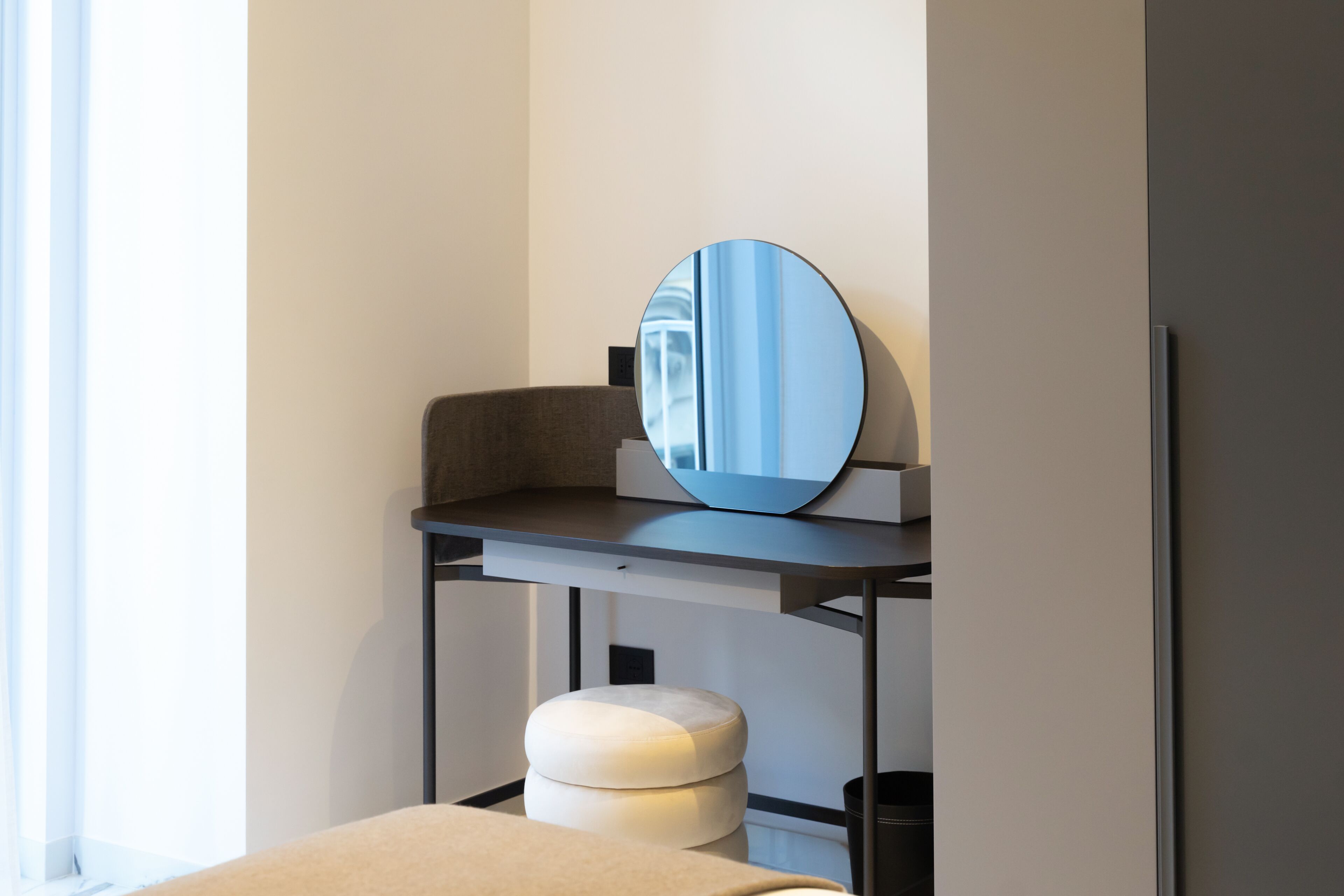 A modern room with a round mirror on a dark console table, a white ottoman, and a black trash bin.