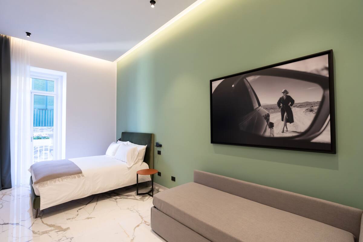 A modern hotel room with a bed, a sofa, a framed picture on the wall, and a window with curtains.