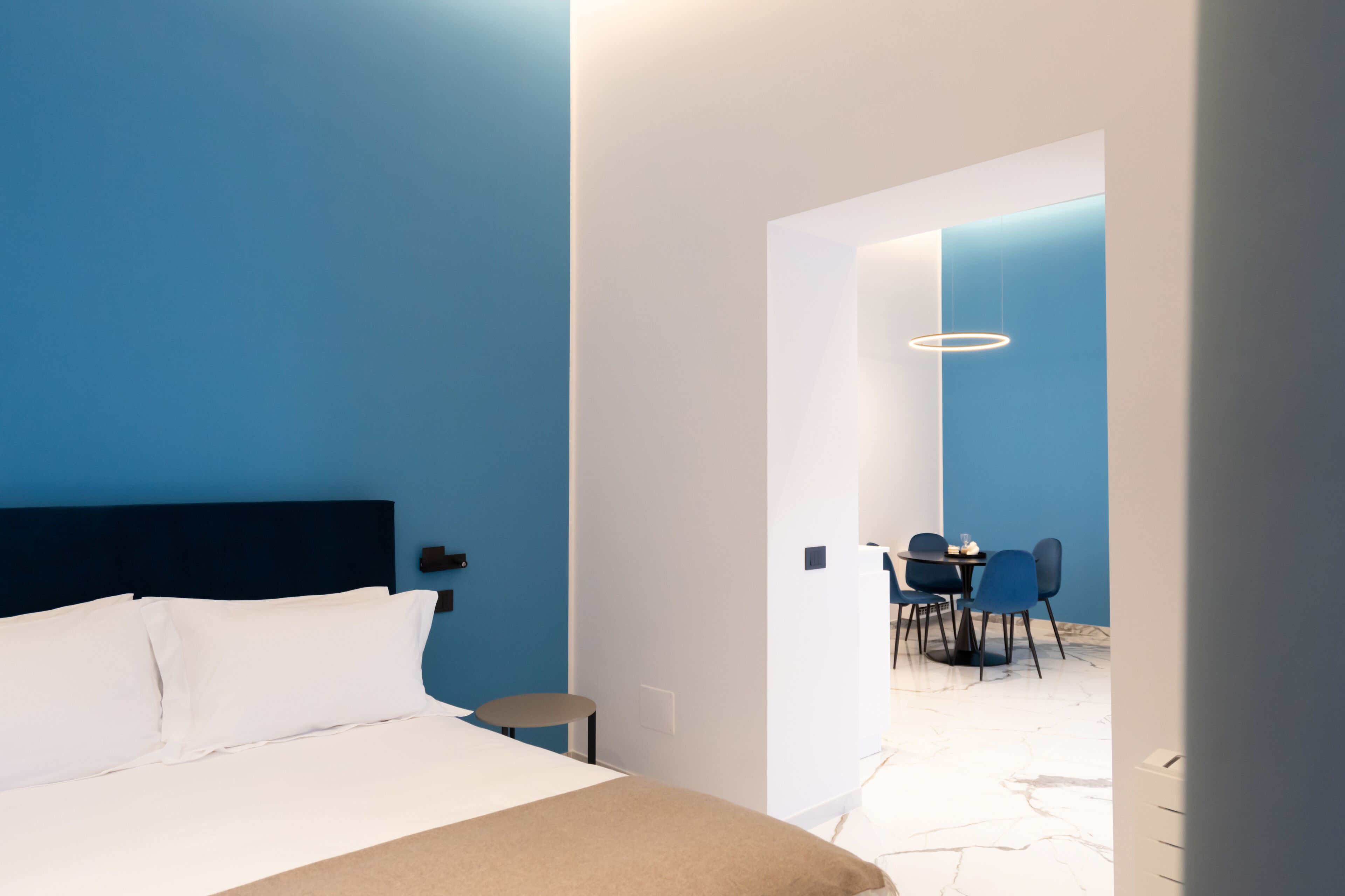 Moderni hotellihuone, jossa on sänky, pieni pöytä ja tuoleja viereisessä ruokailutilassa.