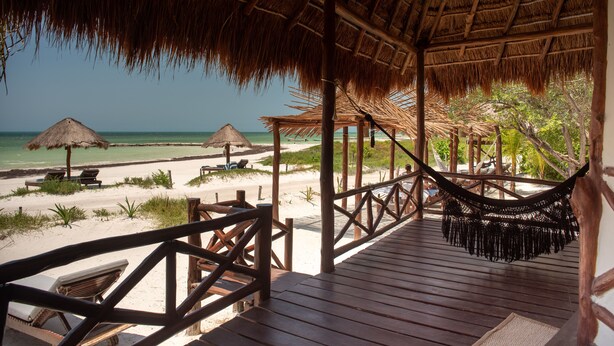 Eine Holzterrasse mit einem Reetdach, mit Blick auf einen Strand mit Liegestühlen und Sonnenschirmen.
