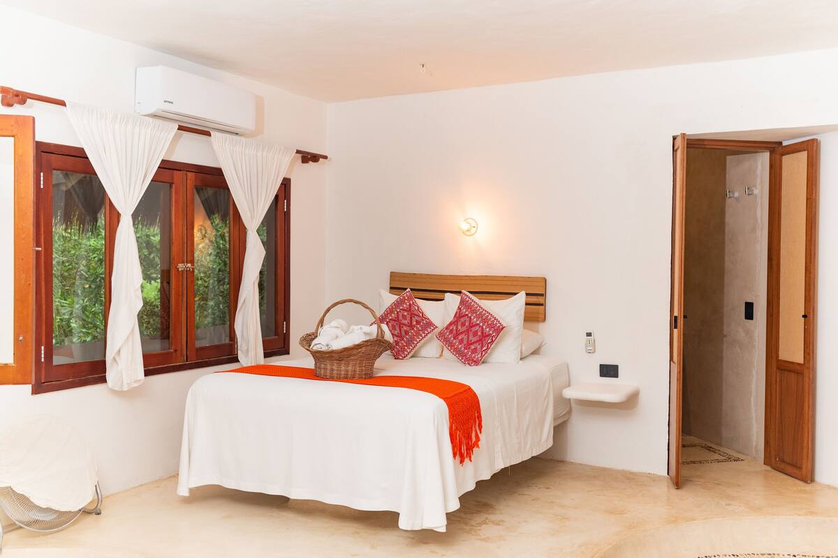 A bedroom with a bed, a bedside table, a wall-mounted air conditioner, a window with curtains, and a door leading to another room.