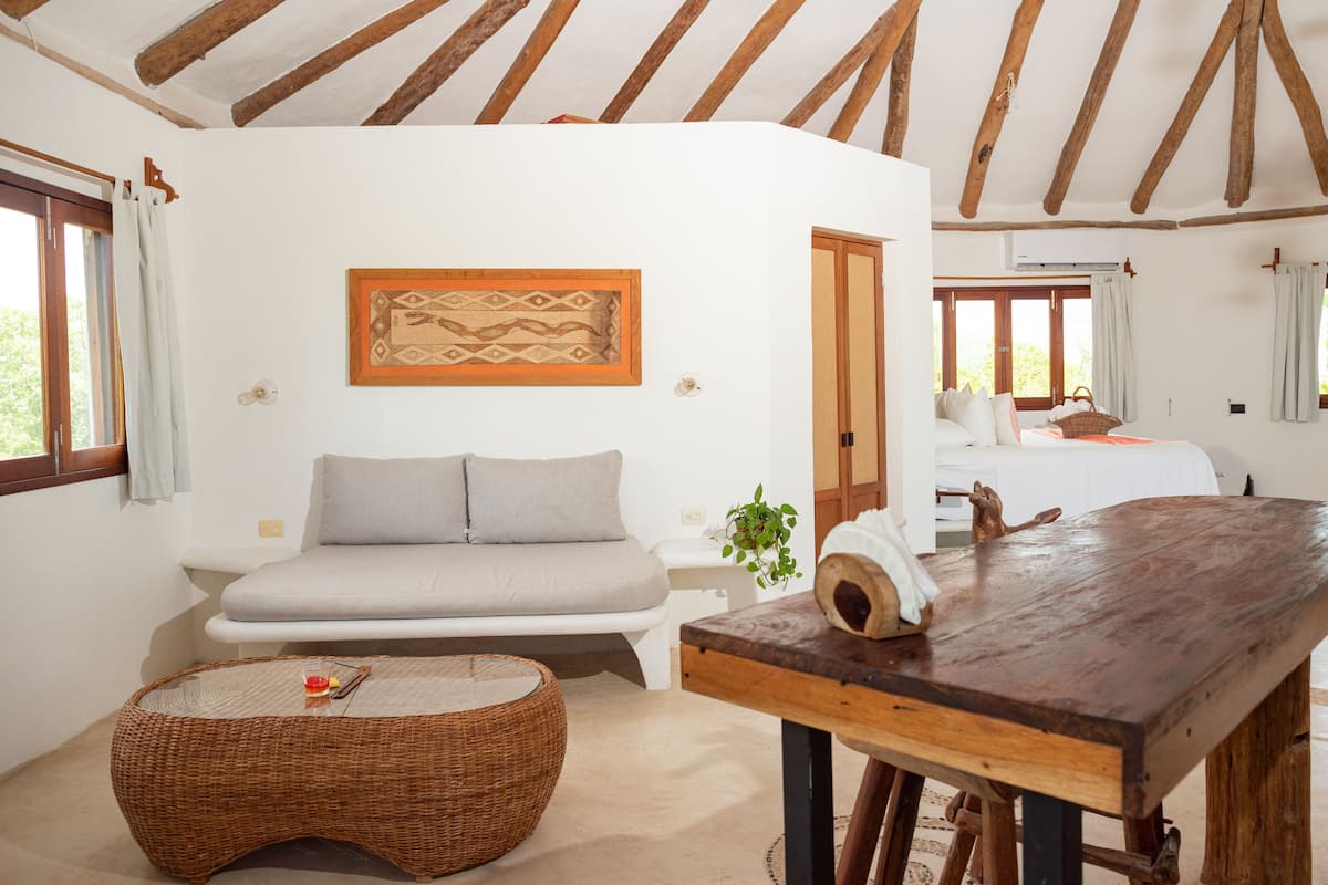 A cozy room with a wooden table, a wicker ottoman, a sofa, and a bed.