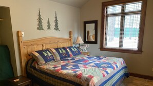 A bedroom with a wooden bed, a nightstand, a chair, a window with blinds, and a wall with three tree decorations. - Quiet apt-cabin on Deschutes River (Bend)