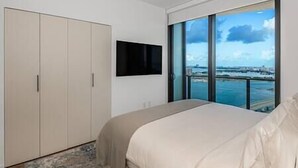 A bedroom with a large bed, a wardrobe, a flat-screen TV mounted on the wall, and a view of a cityscape through large windows.