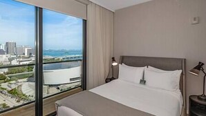 A hotel room with a large window offering a city view, a bed with white linens, and a nightstand with a lamp.