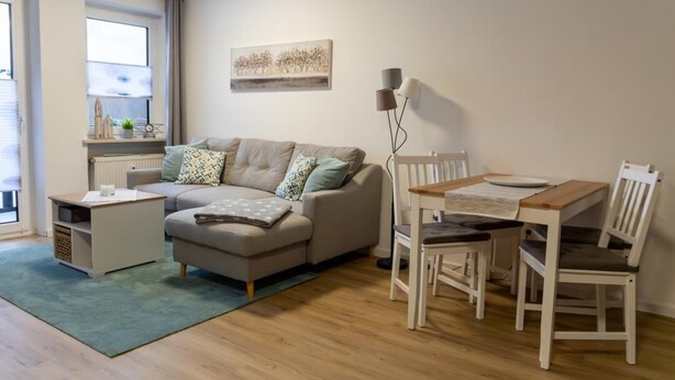 Un salon comprenant un canapé, une petite table, une table à manger avec des chaises, une lampe et une fenêtre avec des stores.