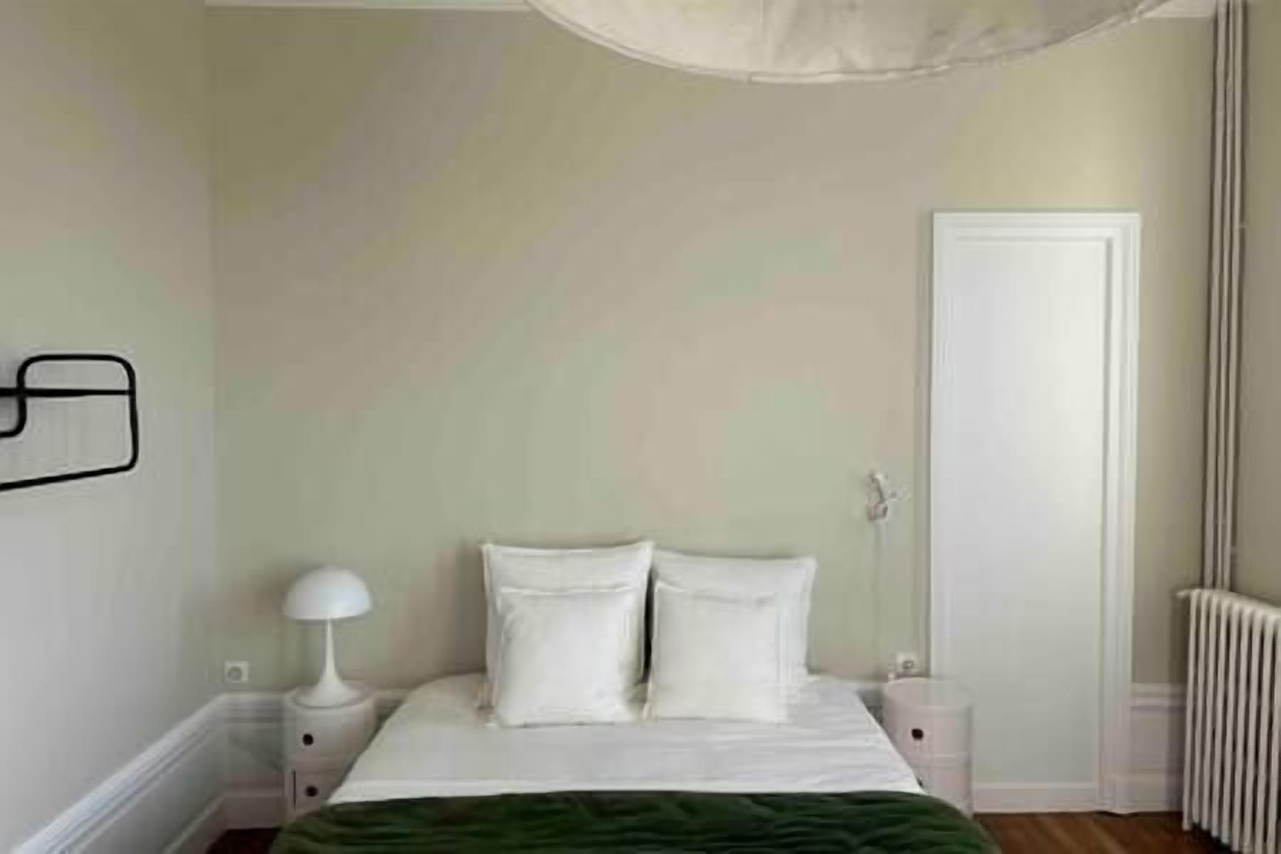 A bedroom with a bed, white walls, a door, a radiator, and a nightstand with a lamp.