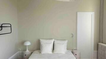 A bedroom with a bed, white walls, a door, a radiator, and a nightstand with a lamp.