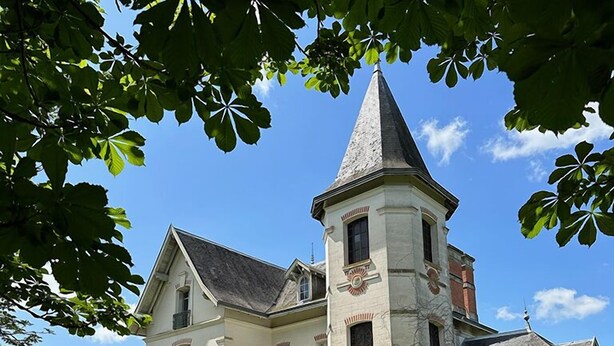 Un edificio grande e histórico con una torre destacada, rodeado de exuberante vegetación y un césped cuidado.