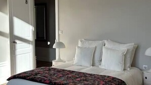 Een slaapkamer met een bed, witte kussens, een bloemenbedekking, een nachtkastje met een lamp, en een deur naar een andere kamer.