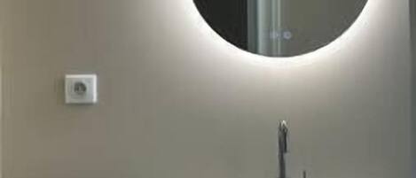 Une salle de bain avec un miroir rond, un lavabo blanc et un interrupteur visible sur le mur.