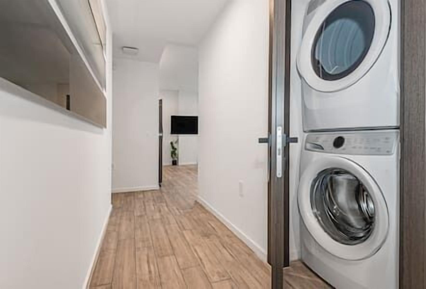 A laundry room with a washing machine and dryer, wooden flooring, and a hallway leading to a living area with a flat-screen TV.