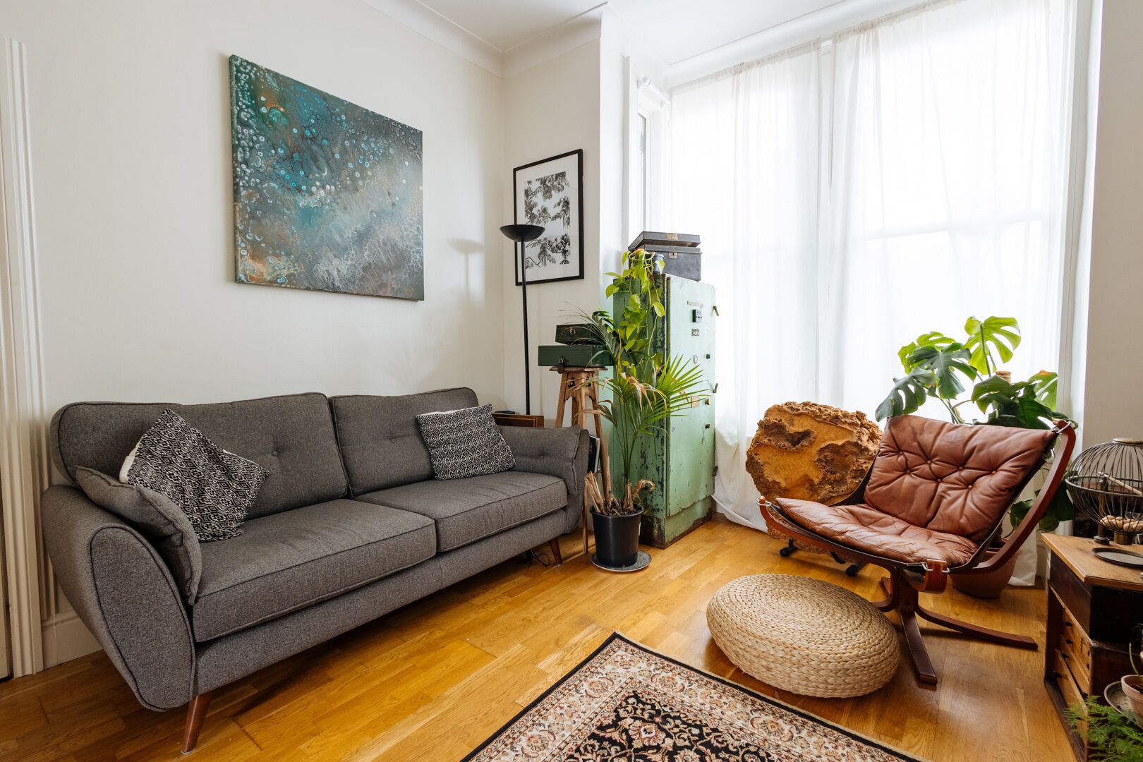 A living room with a grey sofa, a brown leather armchair, a large abstract painting, and a potted plant.