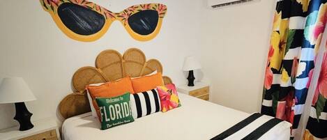 A bedroom with a bed, a ceiling fan, a wall-mounted air conditioner, and a floral curtain.