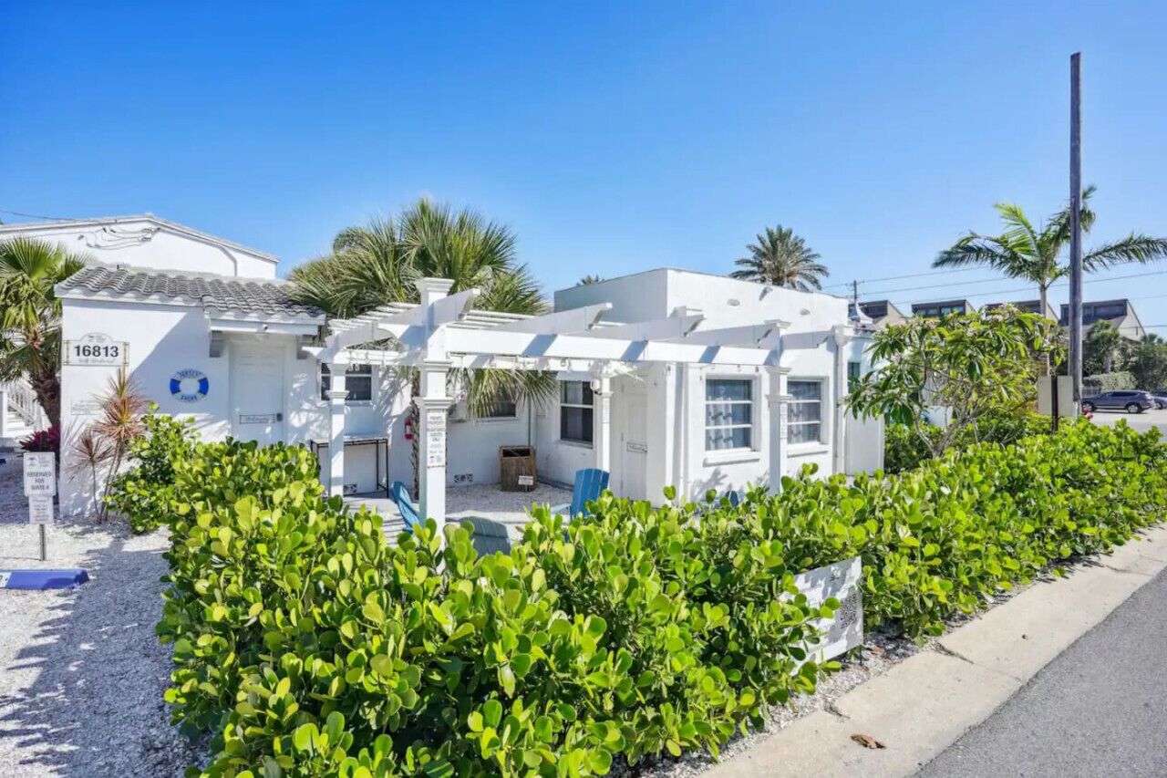 A white building with a tiled roof, surrounded by green shrubs and palm trees.