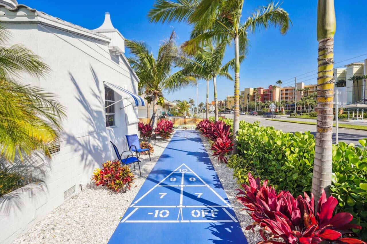 A blue shuffleboard court with numbers, a white building, palm trees, and red plants.