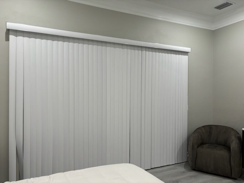 Un dormitorio con un ventanal grande cubierto por persianas blancas, una cama y un sillón oscuro.