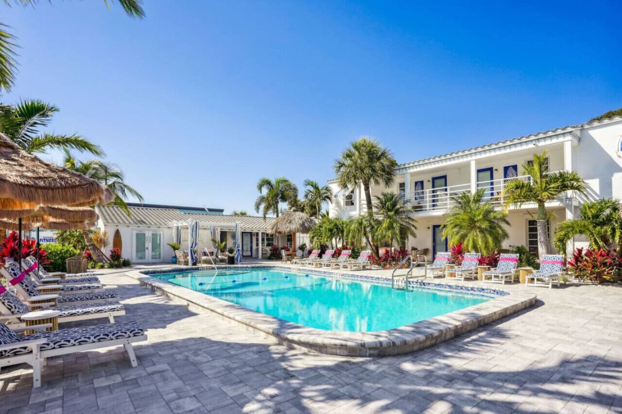 Una piscina con sillas de descanso, palmeras y un edificio de fachada blanca.