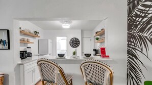 Une cuisine moderne avec des meubles blancs, un îlot central, deux chaises en osier, une horloge murale et une plante décorative.