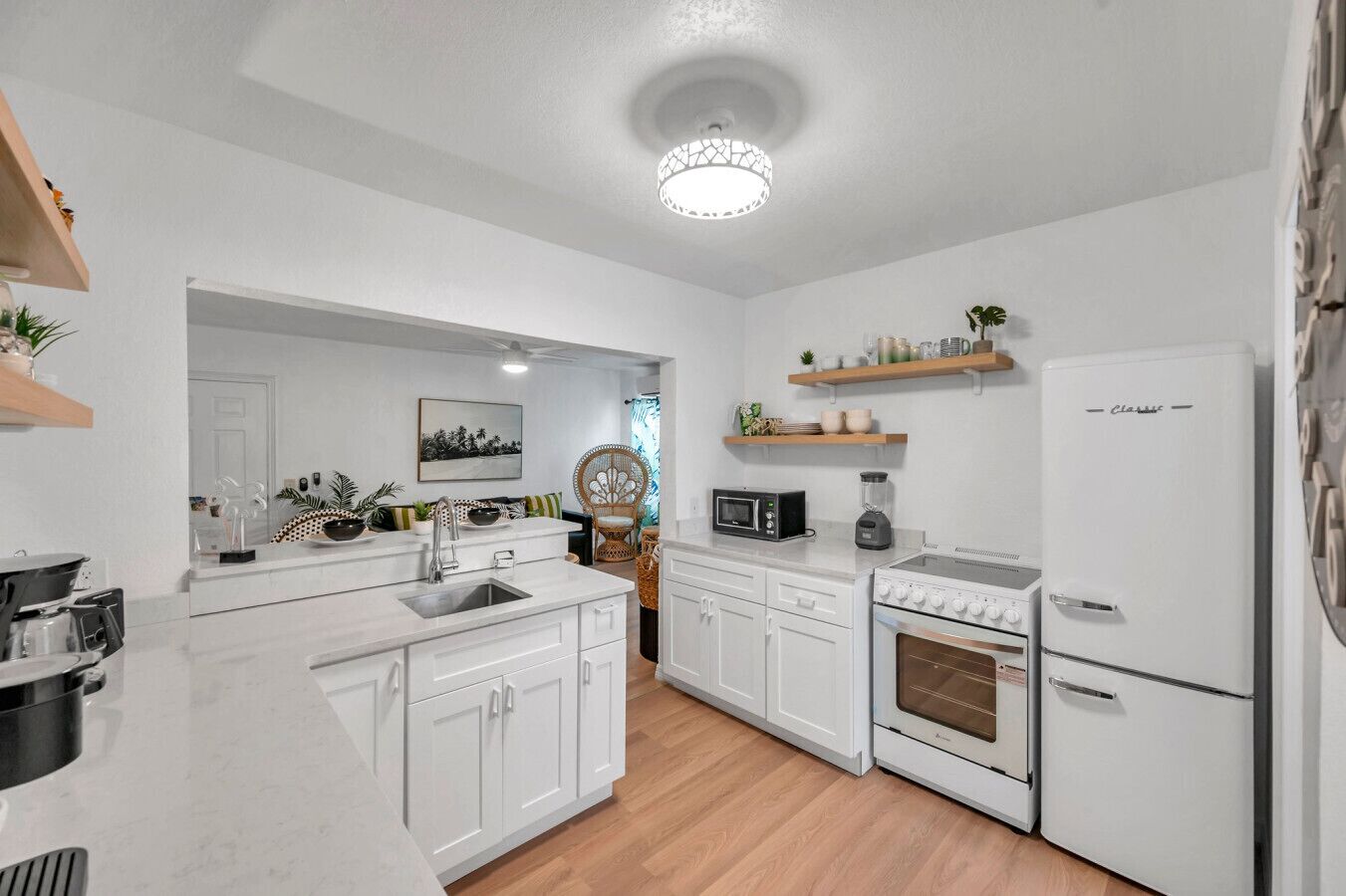 Una cocina moderna con armarios blancos, horno empotrado, microondas y refrigerador. Hay estantes de madera sobre la encimera y una zona de comedor con una silla y una mesa.