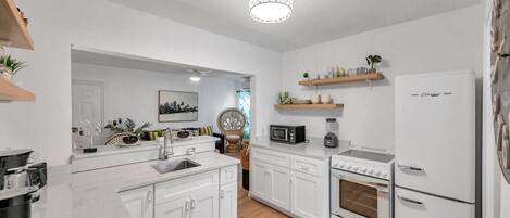 Una cocina moderna con armarios blancos, horno empotrado, microondas y refrigerador. Hay estantes de madera sobre la encimera y una zona de comedor con una silla y una mesa.