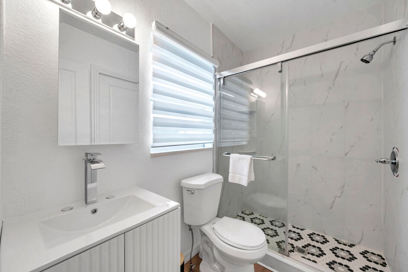 Une salle de bain moderne avec un meuble-lavabo blanc, des toilettes, une douche avec une porte en verre et une fenêtre équipée de stores.